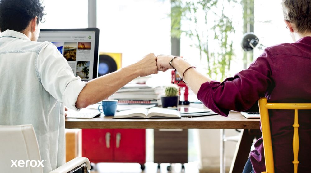 Deux personnes assises à un bureau, dos à la caméra, se serrent le poing, symbole du travail d'équipe et d'une collaboration réussie. L'une d'elles regarde un écran d'ordinateur.